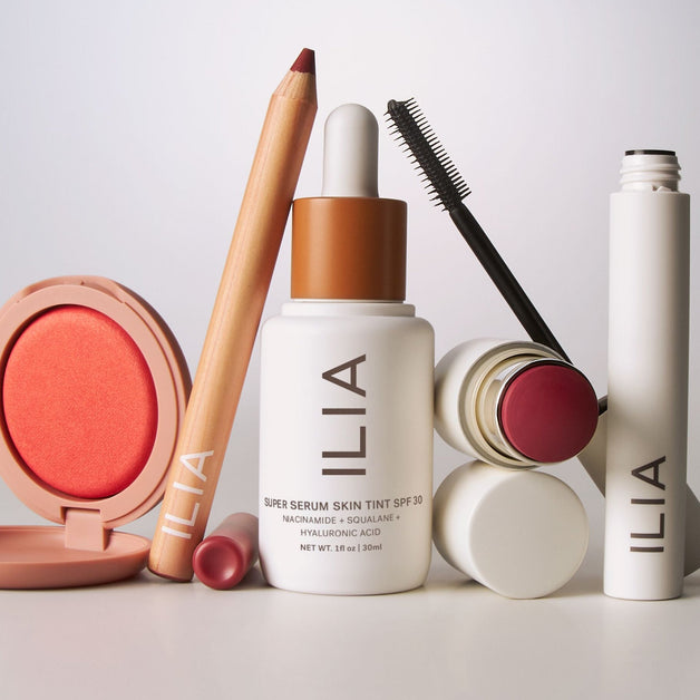 ILIA beauty products arranged on a neutral surface, including mascara, face milk, foundation, and a multi-stick with flowers in the background.
