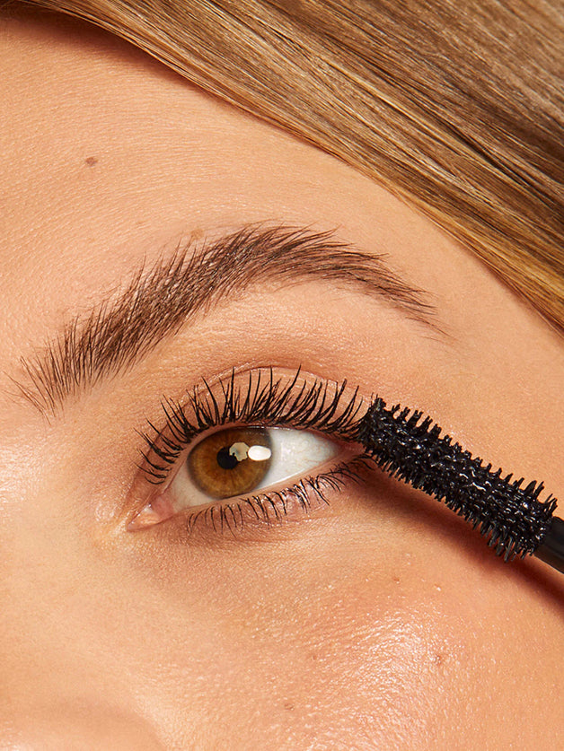 Close-up of a person applying black mascara to their eyelashes, highlighting defined lashes with brown eyes..