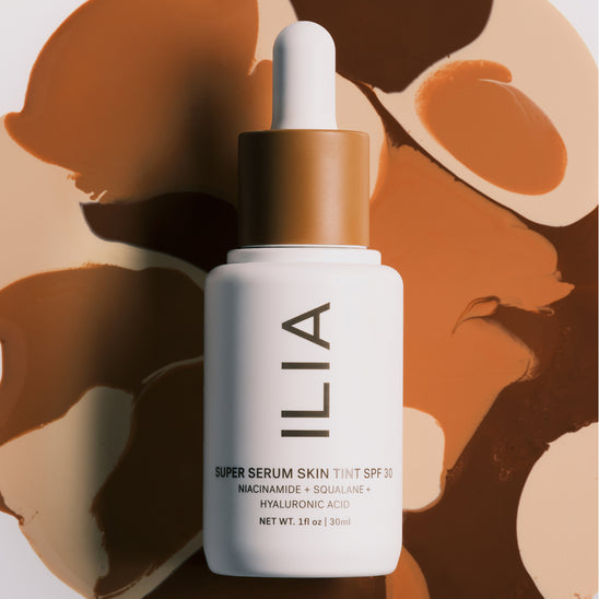 ILIA's Super Serum Skin Tint SPF 30 bottle laid on top of a swirl of different Skin Tint serum shades in the background.
