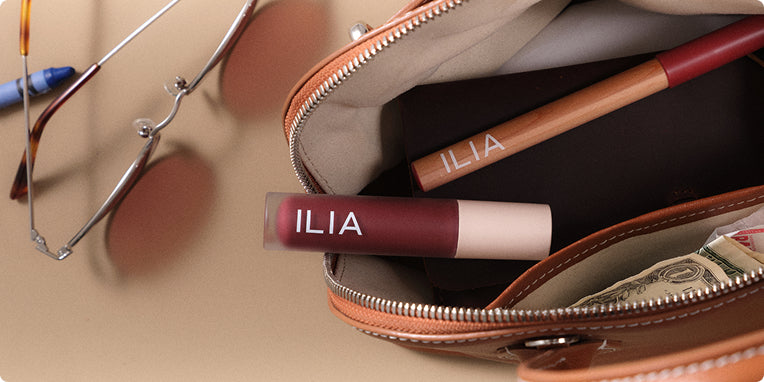 Open handbag with ILIA lip liner, lip gloss and sunglasses on a beige surface